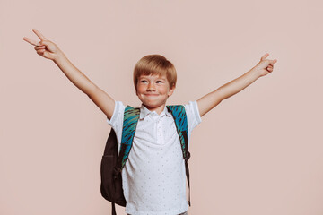School boy with backpack, does peace victory sign with both hands, at studio isolated over beige background. Childhood education concept.