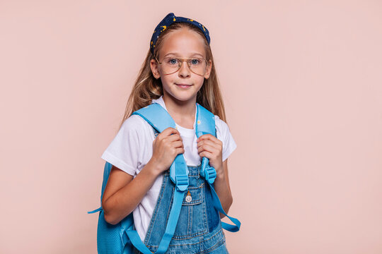 Cute school girl with blue eyes, wears a backpack, eyeglasses and denim overalls, posing at studio isolated over beige background. Childhood education concept.