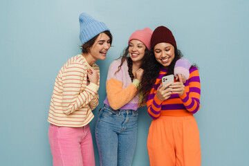 Three cheery girls laughing and using smartphone while standing isolated over blue background