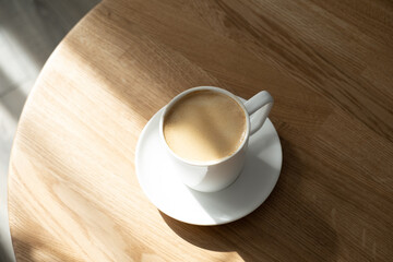A white porcelain cup with coffee on a saucer stands on a small round table made of natural wood. Morning, coffee, hot drink. Hard light.