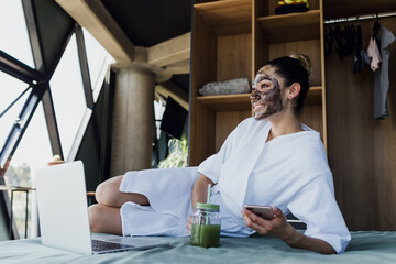 young latin woman with carbon facial mask on her face for Skin care with laptop on bed at home in Mexico, Hispanic female