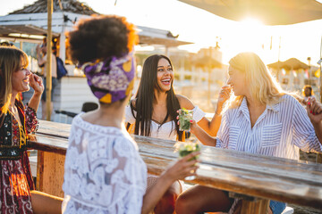 four women having fun at the beach bar, young female friends laughing and chatting, having some drinks and spend time on vacation, holiday and summer concept