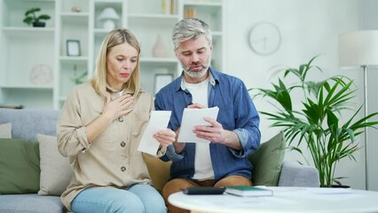 Unhappy shocked married couple looking at large utility bills sitting on sofa at home. Mature sad wife holds a payment check in her hands, and her husband writes down the data in a notebook with a pen - Powered by Adobe