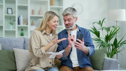 Happy married couple dealing with family finances using online bill payment app while sitting on sofa at home. Mature smiling wife holds checks in her hands and husband records data on smartphone - Powered by Adobe