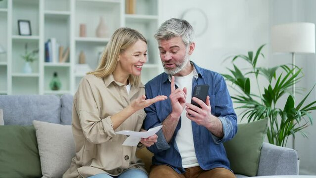 Happy married couple dealing with family finances using online bill payment app while sitting on sofa at home. Mature smiling wife holds checks in her hands and husband records data on smartphone