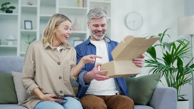 An excited married couple of a customer opens a package at home. Happy husband and wife buyers unpacking purchase sitting on sofa. Satisfied clients of e-commerce. Express delivery services concept