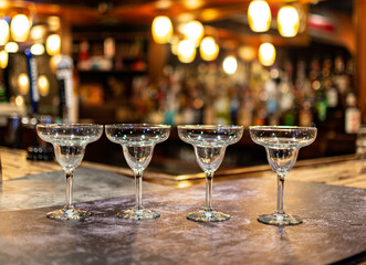 Four long stemmed glasses in focus with bar out of focus in the back. Coupe glasses are upright. The room is glowing with golden bokeh and a party vibe. Moody and Dramatic. 
