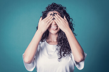 Shot of happy confident curly woman with toothy smile, wears casual basic solid white t-shirt, expresses good emotions, enjoys nice day, covering her face making tongue. Expressions