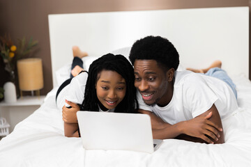 Fototapeta premium Curious surprised dark skinned couple lying on bed in front of laptop smiling eatching comedy movie film. Happy family concept.