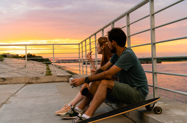 Side view of cheerful young couple resting on embankment against sunset sky and river while eating takeaway food and looking away 