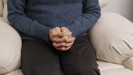 Asian mature woman feels nervous anxious or lonely put clenched hands on laps sitting on couch, sitting on sofa.