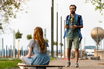 Blurred positive young man with takeaway coffee standing against girlfriend sitting on longboard. Focus at the woman.