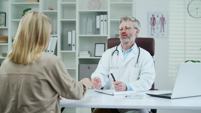 A mature doctor in a white coat consults a patient while sitting at a workplace in a hospital clinic. A male medical worker physician in glasses greets a female visitor and asks about her illness
