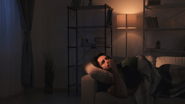 Restless night. Insomnia problem. Mental overload. Sad depressed pensive man overthinking lying on couch late at dark home interior.
