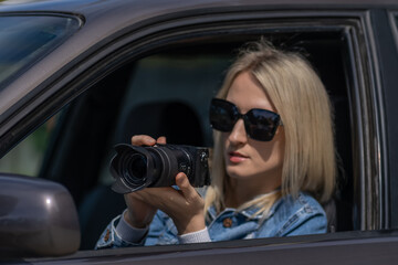 Fototapeta na wymiar Young blonde woman in sunglasses takes pictures on a professional camera while sitting in a car. Concept of journalism, detective, papparation