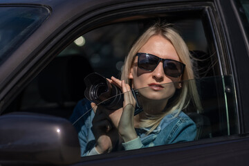 Fototapeta na wymiar Woman in sunglasses with a camera sits in a car and takes pictures with a professional camera, a private detective or a paparazzi spy. Journalist is looking for sensations and follows celebrities.