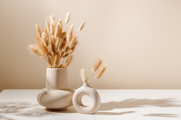Stylish ceramic vase set on the table with dried lagurus grass bouquet with shadows, light brown wall background. Scandinavian vase on the table with copy space, minimal aesthetic interior decoration - obrazy, fototapety, plakaty