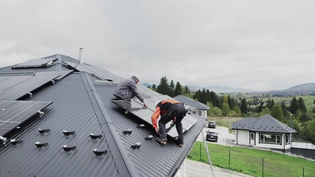Technicians building photovoltaic solar module station on roof of house. Men electricians in helmets installing solar panel system outdoors. Concept of alternative and renewable energy. Aerial view.