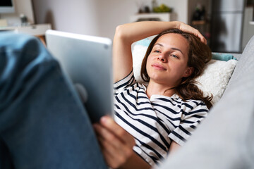 Young woman uses her digital tablet while relaxing at home lying on sofa.