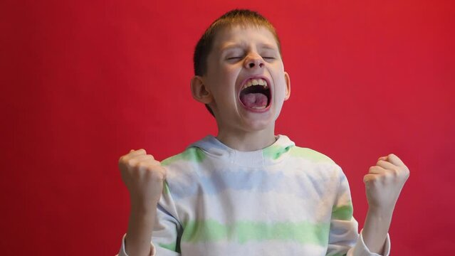happy handsome european boy 9 years old celebrating success raising his hands up saying yes. studio shot, red background. Victory celebration, good luck