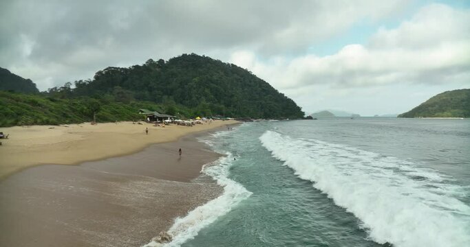 Aerial flying over Ubatuba beach on partly cloudy day, people enjoying vacation