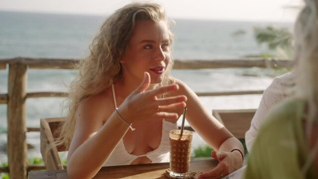 Female friends in sea view rooftop tropical restaurant chat cheerfuly, sip cocktails. Blonde caucasian women sit at the table in an outdoor ocean view cafe talk, smile. Girls at seaside bar