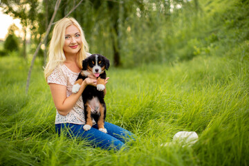 Naklejka na ściany i meble A beautiful girl with a dog sits in the park on the grass. Love to the animals. Home pet on a walk. Animal protection. Bernese mountain dog puppy.