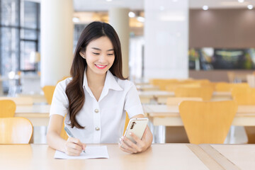 Young Asian student girl in uniform using smartphone and writing something about work. Her face with smiling in working at to search information for study at the university reading room.