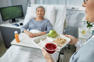 Close-up of nurse carrying tray with healthy food for sick patient lying on bed in hospital ward