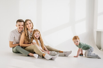 Funny parents hug children sitting on floor at home. Childs play toys, girl and boy. Family moving into new house. Relocation concept. Young happy mother, father, daughter, son isolated on white wall.