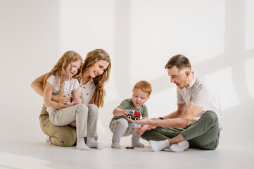 Funny parents hug children sitting on floor at home. Childs play toys, girl and boy. Family moving into new house. Relocation concept. Young happy mother, father, daughter, son isolated on white wall.