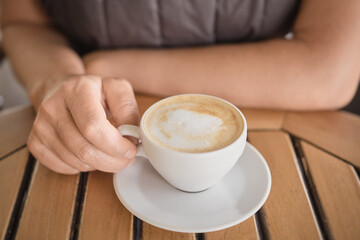 Close-up of a cup of fragrant cappuccino on the table in the hands of a woman, invigorating fragrant coffee during a break. Selective focus on fingers, idea for background, natural sunlight