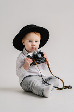 Child in a hat sitting on floor and play with photo camera. Fun kid holds retro vintage camera isolated on white wall. International Photographers Day. Children's studio portrait. Mockup. Close Up.