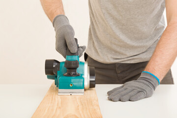 Young adult man hands using electric jointer and shaving old wooden plank for furniture or floor on white table at light gray wall. Closeup. Preparing material for repair work of home. Front view.