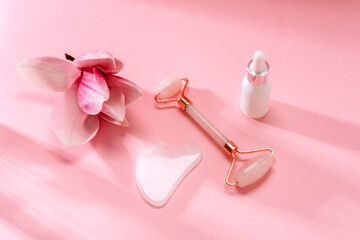 Obraz na płótnie Canvas Rose quartz crystal facial roller and gua sha scraper, face serum or organic cosmetic oil, magnolia flower on pink background with long shadows. Facial massage kit for lifting therapy. Top view