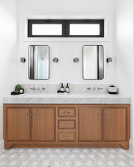 A cozy bathroom with a wood cabinet, marble countertop, sconces around square mirrors, and a beautiful tiled floor.