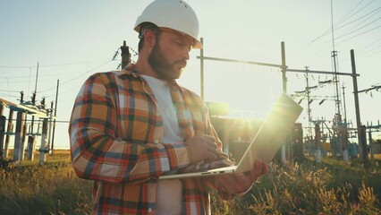 Design engineer in helmet holds laptop in hands at construction site in field, estimating investment attractiveness this object. Male compares his project with customer project. Using technologies.