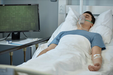 Young man lying on bed with oxygen mask on his face during treatment in hospital ward