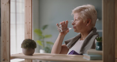 Senior caucasian woman drinking water agter finishing her training session. Mature athlete keeping herself hydrated after yoga - healthy lifestyle, wellbeing 