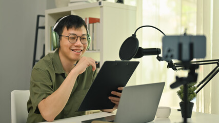 Smiling young man radio host wearing headphone streaming his live podcast using professional...