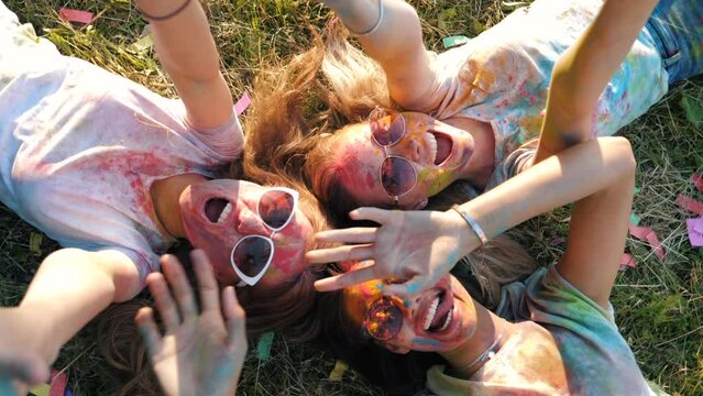 Three happy beautiful female making party at Holi colours festival in summer time. Young  smiling women friends having fun after music event at sunset. Models lying on the grass in sunglasses