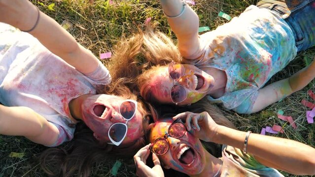 Three happy beautiful female making party at Holi colours festival in summer time. Young  smiling women friends having fun after music event at sunset. Models lying on the grass in sunglasses