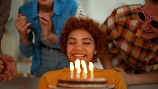 African american girl celebrates birthday with friends blows candles on cake makes a wish. They smile and clap hands. Wearing festive caps and blows party horns. Celebration party for birthday girl.