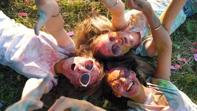 Three happy beautiful female making party at Holi colours festival in summer time.Young  smiling women friends having fun after music event at sunset.Models lying on the grass in sunglasses