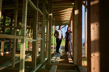 Fototapeta premium Man and woman inspecting their future wooden frame house nestled near the forest. Youthful couple at construction site in early morning. Concept of contemporary ecological construction.