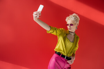 Fototapeta na wymiar tattooed albino woman in stylish sunglasses taking selfie on mobile phone on carmine pink background.