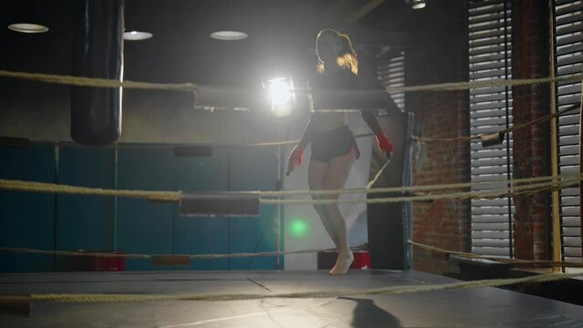Professional young sportswoman boxer in sportswear with ponytail skipping rope on boxing ring in gym. She training alone. Girl jumping rope. Sport, workout, train, fighter training, jump rope concept.
