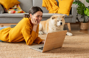 Smiling woman lying on carpet and using laptop by dog at home