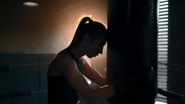 Silhouette of young tired exhausted sportswoman boxer loaning head and hands to punching bag in dark, side view. Sweaty girl is resting after hard training. Workout, box, professional sport concept.