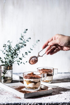 Hand of woman sprinkling cocoa powder through colander on tiramisu in mug